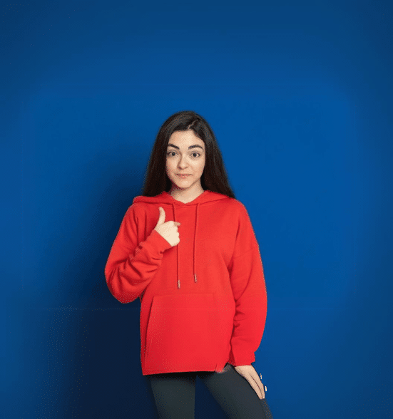 Young woman in a red hoodie giving a thumbs-up gesture with a confident expression against a blue background.