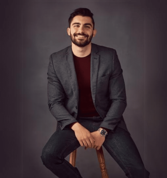 Confident young man in a blazer sitting on a stool, smiling, with a stylish and professional appearance.